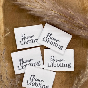 May include: Five white fabric labels with black text that reads "kleiner Liebling" which translates to "little darling" in English. The labels are on a wooden surface with a dried grass sprig.