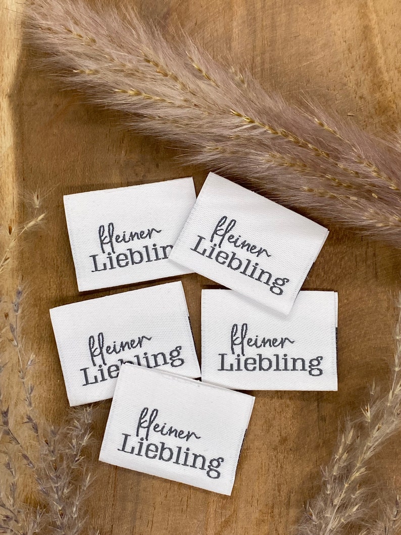 May include: Five white fabric labels with black text that reads "kleiner Liebling" which translates to "little darling" in English. The labels are on a wooden surface with a dried grass sprig.