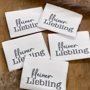 May include: Four white fabric labels with black embroidery that reads "kleiner Liebling" which translates to "little darling" in English.