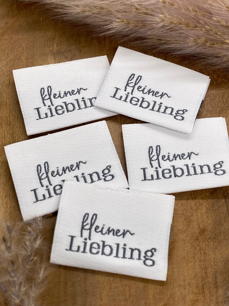 May include: Four white fabric labels with black embroidery that reads "kleiner Liebling" which translates to "little darling" in English.