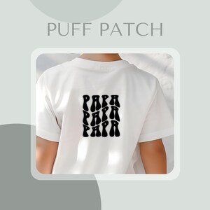 May include: White t-shirt with a black "PAPA" puff patch design on the back. The text is in a retro, wavy font. The shirt is short-sleeved and appears to be made of a soft material. The words "PUFF PATCH" are at the top.