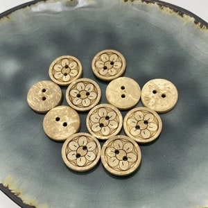 May include: A selection of beige wooden buttons, some with two holes, others with four, and some with a carved floral pattern. They are arranged on a teal ceramic plate with a dark brown rim, creating a natural, earthy aesthetic.