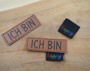 May include: Two brown leather patches with the text "ICH BIN" in black lettering.  A black fabric patch with the text "happy" in rainbow lettering is placed on top of one of the leather patches.