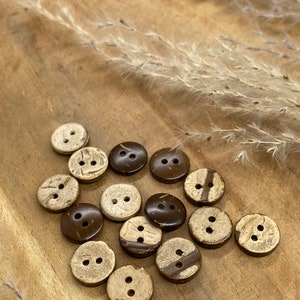 May include: A collection of 14 brown and beige coconut shell buttons with two holes each. The buttons are arranged in a circle on a wooden surface.