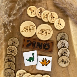 Könnte beinhalten: Holzknöpfe mit Dinosaurier-Designs, darunter ein Triceratops, ein Stegosaurus und ein T-Rex. Die Knöpfe sind auf einer Holzoberfläche mit einem Leder-Patch, auf dem "DINO" steht, und zwei Stoff-Patches mit Dinosaurier-Illustrationen angeordnet.