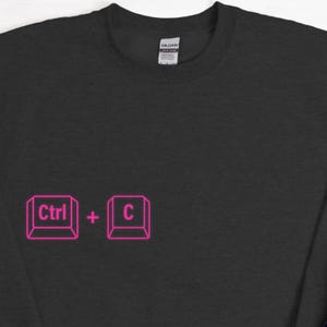 Może przedstawiać: Czarna bluza z okrągłym dekoltem z r&oacute;żową grafiką skr&oacute;t&oacute;w klawiaturowych "Ctrl + C". Bluza wykonana jest z miękkiego materiału i ma klasyczny kr&oacute;j. Etykieta marki Gildan jest widoczna z tyłu szyi.