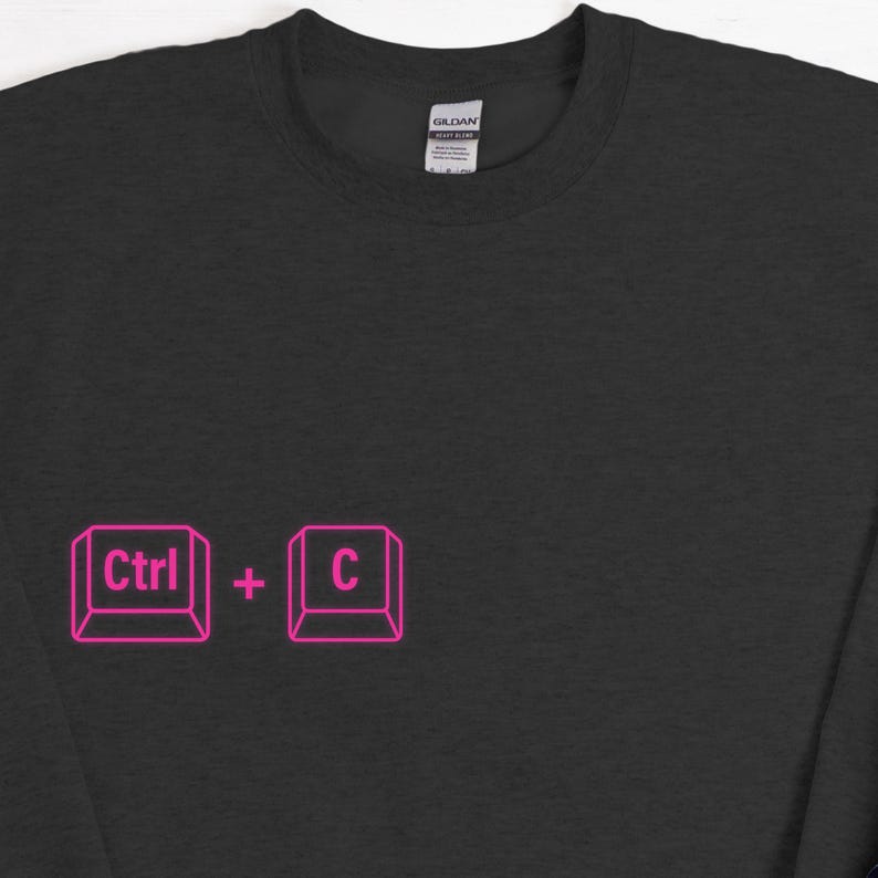 Może przedstawiać: Czarna bluza z okrągłym dekoltem z r&oacute;żową grafiką skr&oacute;t&oacute;w klawiaturowych "Ctrl + C". Bluza wykonana jest z miękkiego materiału i ma klasyczny kr&oacute;j. Etykieta marki Gildan jest widoczna z tyłu szyi.