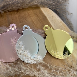 May include: Three round, mirrored Christmas baubles in pink, silver, and gold. The baubles are hanging from a wooden surface.