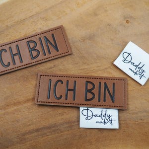 Könnte beinhalten: Zwei braune Leder-Etiketten mit dem Text "ICH BIN" in schwarzer Schrift. Ein weißes Etikett mit dem Text "Daddy made it" in schwarzer Kursivschrift.