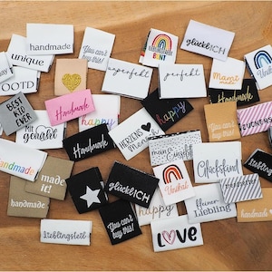 Könnte beinhalten: Eine Sammlung kleiner, rechteckiger Stoffetiketten mit verschiedenen gestickten Phrasen und Designs. Die Etiketten sind in Weiß, Schwarz, Rosa und Beige gehalten und tragen Aufschriften wie "handmade", "happy" und "love". Einige zeigen Regenbogen, Herzen und Sterne.