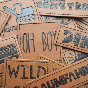 Può includere: Una collezione di toppe in pelle marrone con testo nero e disegni. Le toppe presentano frasi come "Oh Boy", "Wild", "Tatum", "Dino" e "Monster".