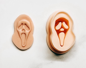 Puede incluir: Dos cortadores de galletas de color beige con forma de la máscara de Ghostface de la franquicia de películas de terror Scream. Un cortador es una forma sólida y el otro es un cortador en capas. Los cortadores están sobre una superficie blanca.