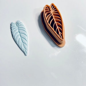 Puede incluir: Dos cortadores de galletas con forma de hoja, uno azul claro y otro marr&oacute;n. El cortador marr&oacute;n es un dise&ntilde;o de doble cara con un contorno elevado y un centro recortado.