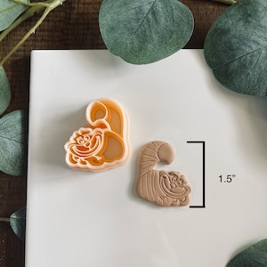 May include: Two cookie cutters, one in light orange and one in brown, both shaped like the Cheshire Cat from Alice in Wonderland. The brown cutter is 1.5 inches tall.