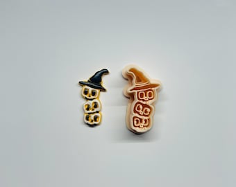 Puede incluir: Dos adornos de Halloween. Uno es negro, dorado y blanco, el otro es marrón y beige. Ambos presentan una pila de tres caras sonrientes con la palabra "BOO" y un sombrero de bruja. Probablemente para manualidades o decoración de temporada.