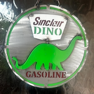 May include: Circular metal sign featuring a green dinosaur silhouette within a green-bordered circle. The sign reads "Sinclair DINO GASOLINE" in white and red lettering. The sign is suspended by a chain.