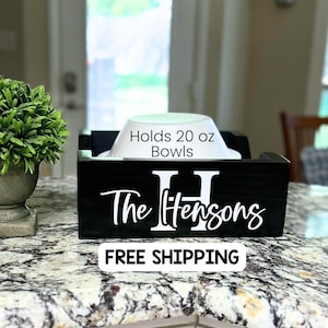 May include: A black wooden bowl holder with a white bowl inside. The holder has the text "The Hensons" and the letter "H" in white. The text "Holds 20 oz Bowls" is also visible.