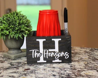 Könnte beinhalten: Ein dunkel gebeizter Holz-Schreibtisch-Organizer mit dem Namen "The Hensons" und einem großen weißen "H" auf der Vorderseite. Ein roter Becher und ein schwarzer Marker befinden sich im Inneren. Links ist eine kleine grüne Pflanze in einem grauen Topf.