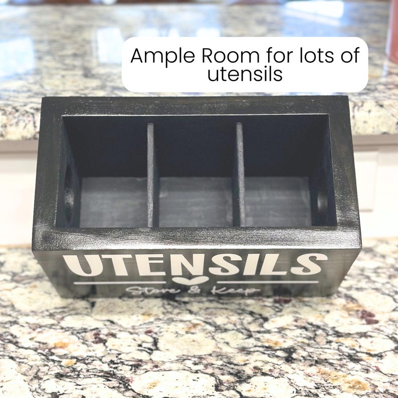 Op de afbeelding: Een zwarte houten keukengerei houder met drie compartimenten. De houder heeft het woord "UTENSILS" in witte letters op de voorkant, samen met de zin "Store & Keep". De bovenkant van de houder heeft de tekst "Ample Room for lots of utensils".