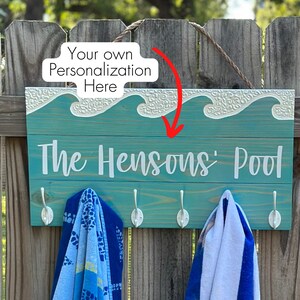 K&ouml;nnte beinhalten: Ein t&uuml;rkisfarbenes Holzschild mit wei&szlig;er Aufschrift "The Hensons' Pool". Das Schild hat ein dekoratives Wellendesign und Haken f&uuml;r Handt&uuml;cher. Das Schild h&auml;ngt an einem Holzzaun.