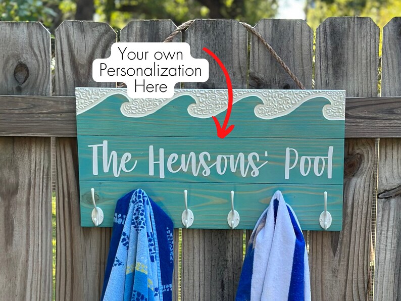 K&ouml;nnte beinhalten: Ein t&uuml;rkisfarbenes Holzschild mit wei&szlig;er Aufschrift "The Hensons' Pool". Das Schild hat ein dekoratives Wellendesign und Haken f&uuml;r Handt&uuml;cher. Das Schild h&auml;ngt an einem Holzzaun.