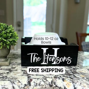 May include: Black wooden bowl holder with the text "The Hensons" and a large white "H". The holder is designed to hold 10-12 oz bowls. A white bowl is placed inside the holder.