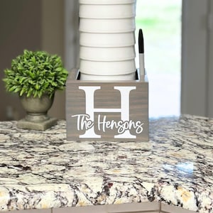 May include: A gray wooden kitchen utensil holder with the letter "H" and the name "The Hensons" in white. The holder contains white bowls and a black marker. A small green plant in a gray pot is to the left.