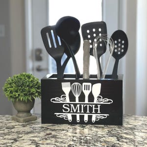 Op de afbeelding: Een zwarte houten keukengerei houder met de naam "SMITH" in witte letters. De houder bevat diverse zwarte keukengereedschappen, waaronder een sleuflepel, spatel en garde. Links staat een kleine groene plant in een grijze pot.