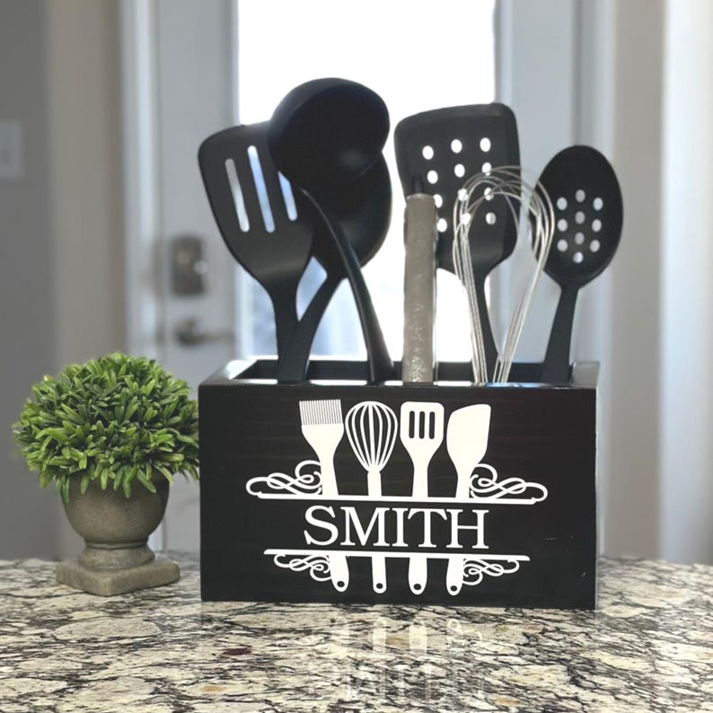 Op de afbeelding: Een zwarte houten keukengerei houder met de naam "SMITH" in witte letters. De houder bevat diverse zwarte keukengereedschappen, waaronder een sleuflepel, spatel en garde. Links staat een kleine groene plant in een grijze pot.