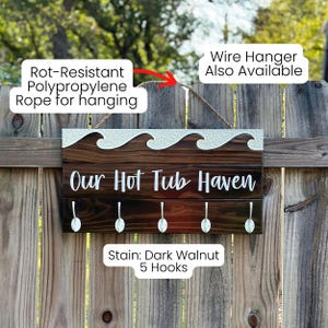 K&ouml;nnte beinhalten: Dunkel walnussfarbenes Holzschild mit dem Text "Our Hot Tub Haven" und f&uuml;nf Haken. Das Schild hat ein Wellendesign und ein Polypropylen-Seil zum Aufh&auml;ngen. Ein dekorativer Artikel.