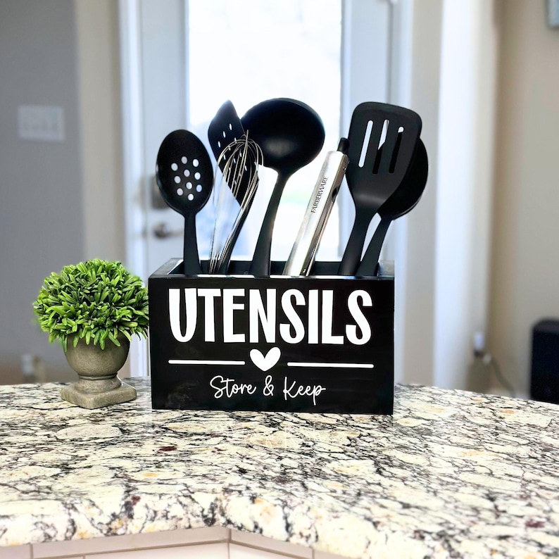 Op de afbeelding: Een zwarte houten keukengerei houder met het woord "UTENSILS" in het wit. De houder bevat zwarte keukengereedschappen, waaronder een sleuflepel, garde, pollepel, spatel en een zilveren keukengerei. De tekst "Store & Keep" staat eronder.