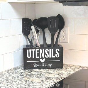 Op de afbeelding: Een zwarte houten keukengerei houder met het woord "UTENSILS" in het wit, een hart en de woorden "Store & Keep". De houder bevat diverse zwarte keukengerei, waaronder een garde, een spatel en een sleef. De houder staat op een aanrecht.