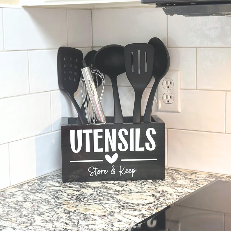 Op de afbeelding: Een zwarte houten keukengerei houder met het woord "UTENSILS" in het wit, een hart en de woorden "Store & Keep". De houder bevat diverse zwarte keukengerei, waaronder een garde, een spatel en een sleef. De houder staat op een aanrecht.