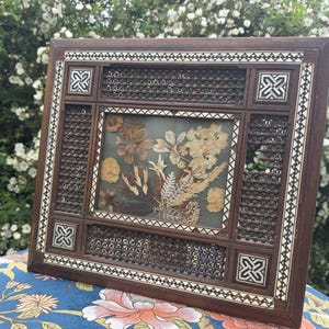 May include: A rectangular wooden frame featuring intricate dark brown lattice panels and inlaid geometric mother-of-pearl patterns. The centre glass display contains a collection of pressed dried flowers and foliage in shades of beige and brown.