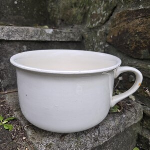May include: A white ceramic chamber pot with a handle. The pot is round and has a smooth, glossy finish.