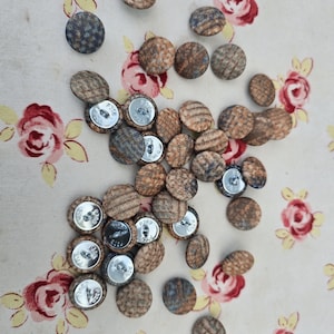 May include: A scattering of round fabric-covered buttons in shades of brown, blue, and grey. The buttons feature textured patterns and silver metal backs. The background is a floral fabric with red and pink roses.