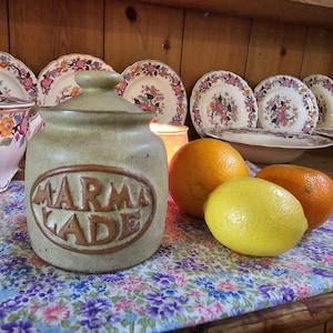 May include: A stoneware marmalade jar with a lid, labelled "MARMALADE" in a brown oval. Two oranges and a lemon sit on a floral patterned cloth. Floral plates and bowls are in the background.