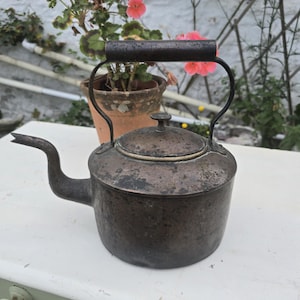 May include: Vintage metal teapot with a curved spout, a lid, and a handle. The teapot has a dark, aged appearance, suggesting it is antique. The background includes a potted plant with pink flowers and a white surface.