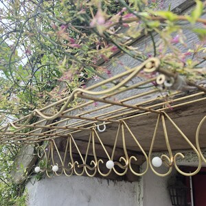 May include: A gold-toned metal wall-mounted rack featuring a rectangular wire shelf top and a series of downward-facing hooks. Each hook is finished with a small, round white bead at the tip, and the metal frame displays a decorative scrollwork design.