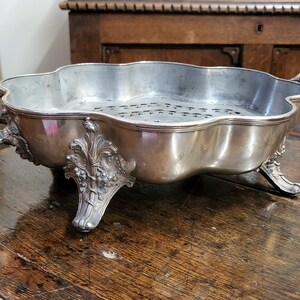 May include: A silver-plated serving dish with a decorative design and three legs. The dish has a perforated bottom and two ornate handles.