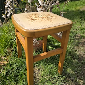 May include: A wooden stool with a square, cushioned seat featuring a floral design in shades of brown and beige. The stool has four sturdy, light brown legs and is set outdoors on a grassy surface.