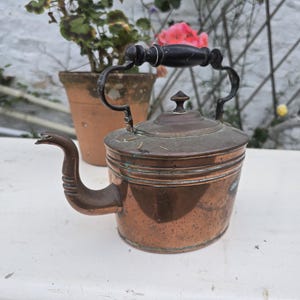 May include: Vintage copper kettle with a dark wooden handle and spout. The kettle has a round body and a lid with a small finial. The handle is arched and attached to the body with metal supports. The kettle is on a white surface.