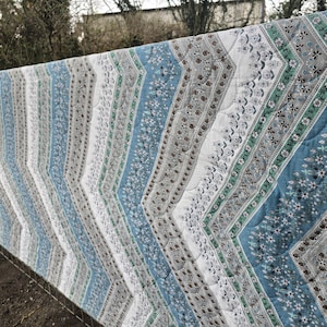 May include: A patterned quilt with a chevron design, displayed outdoors. The quilt combines shades of blue, white, grey, and green, with floral and geometric motifs. The fabric's intricate details are visible, highlighting the quilt's craftsmanship.