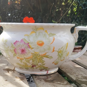 May include: A white ceramic chamber pot with a handle and floral design. The pot features a large, ornate handle and is decorated with pink, yellow, and green flowers. The pot is sitting on a weathered wooden surface.