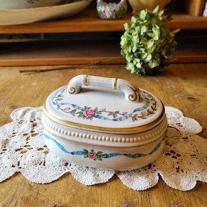 May include: White porcelain oval-shaped trinket box with a gold trim and a blue and white ribbon design. The lid has a gold handle and a pink rose design.