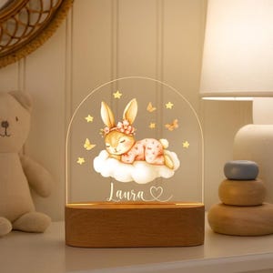 May include: A decorative night light featuring a sleeping bunny on a cloud, with the name "Laura" and a heart. The bunny wears a floral crown and is surrounded by stars and butterflies. The base is made of wood.