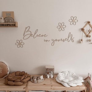 Könnte beinhalten: Ein hellbraunes Holzschild mit den Worten "Believe in yourself" in kursivschrift. Das Schild hängt an einer weißen Wand mit ein paar kleinen Holz-Gänseblümchen-Dekorationen. Es gibt auch ein paar Holz-Aufbewahrungskisten mit Etiketten.