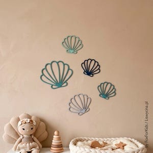 Puede incluir: Cinco decoraciones de pared de conchas marinas recortadas en varios tamaños y tonos de azul, verde azulado y gris. Las conchas presentan un borde festoneado y nervaduras verticales. El texto "by FoxForKids / Liswycina.pl" está impreso.