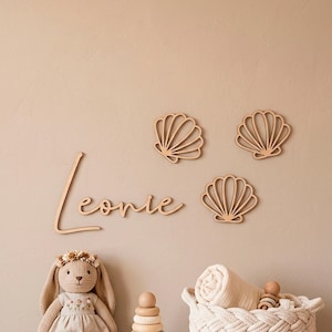 May include: A light brown wooden wall cutout of the name Leonie in cursive script, accompanied by three matching wooden seashell cutouts. Below are a crocheted rabbit doll, a wooden stacking toy, and a woven basket containing a rolled fabric.