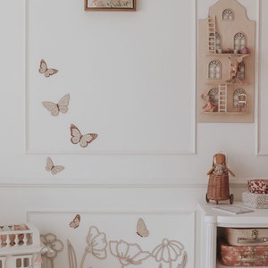 May include: A light-toned nursery scene with wooden butterfly and flower wall decorations. A wooden dollhouse and a decorative house-shaped shelf with miniature figures are also visible. A doll in a wicker cart and stacked suitcases complete the decor.